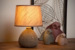 Brązowa lampa stołowa Ramzi z okrągłym, teksturowanym podstawą i kloszem w odcieniu jasnego beżu. Ujęcie frontalne, lampa świeci ciepłym światłem, w tle widoczne dekoracyjne figurki ptaków na drewnianym blacie.
