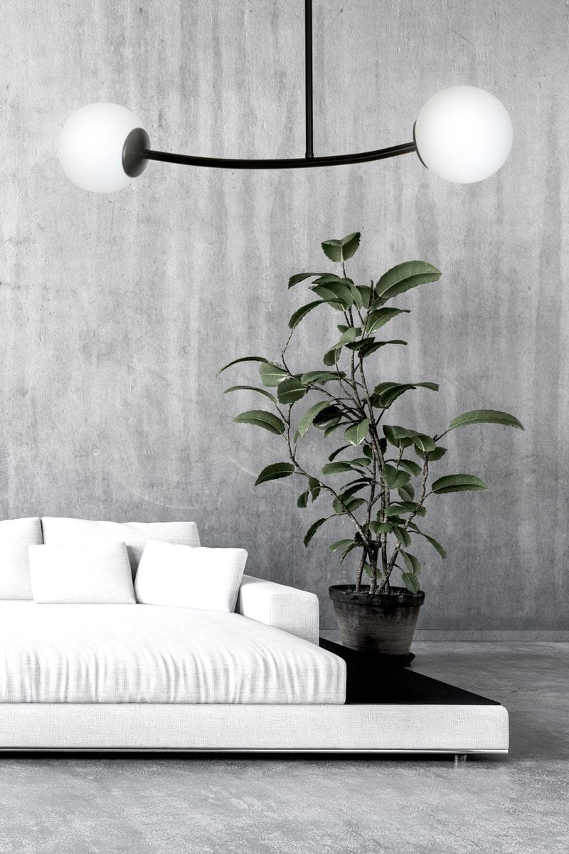 Czarna lampa wisząca Halldor 2-punktowa z białymi kulistymi kloszami, zawieszona na cienkim, łukowatym ramieniu. Produkt w nowoczesnym wnętrzu z szarymi ścianami, obok białej sofy i doniczki z rośliną.