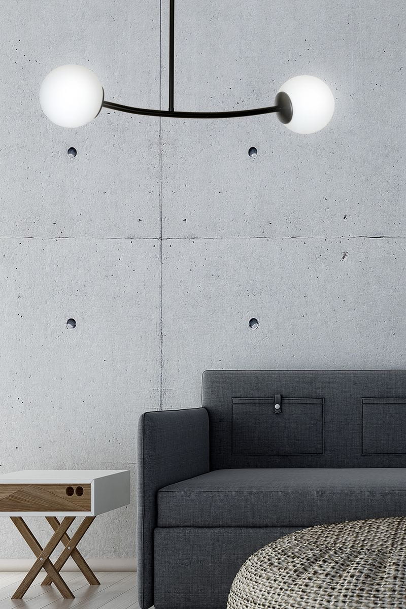 Czarna lampa wisząca Halldor z dwoma okrągłymi kloszami w białym kolorze, umieszczona na cienkim, zakrzywionym ramieniu. Lampa wisi nad szarym sofą w minimalistycznym wnętrzu z betonową ścianą.