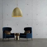 Złota lampa wisząca Telma 1-punktowa o zaokrąglonym kloszu z prążkowaną fakturą, zawieszona nad okrągłym stolikiem w nowoczesnym wnętrzu z szarym tłem. Dwa granatowe fotele z poduszkami w kolorze brązowym tworzą elegancką aranżację.