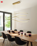Lampa wisząca Waves LED 4-punktowa w kolorze złotym z falistymi elementami, zawieszona na cienkich linkach. Zainstalowana nad drewnianym stołem, w otoczeniu czarnych krzeseł i kolorowych misek. Jasne, nowoczesne wnętrze z dużymi oknami.