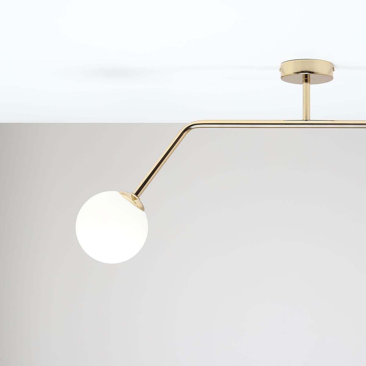 Lampa sufitowa Pure 2-punktowa w kolorze złotym, z dwoma okrągłymi kloszami, umieszczona na długim, zakrzywionym ramieniu. Klosze mają białą, matową powierzchnię, a lampa jest zamontowana na suficie. Tło w odcieniu jasnoszarym.