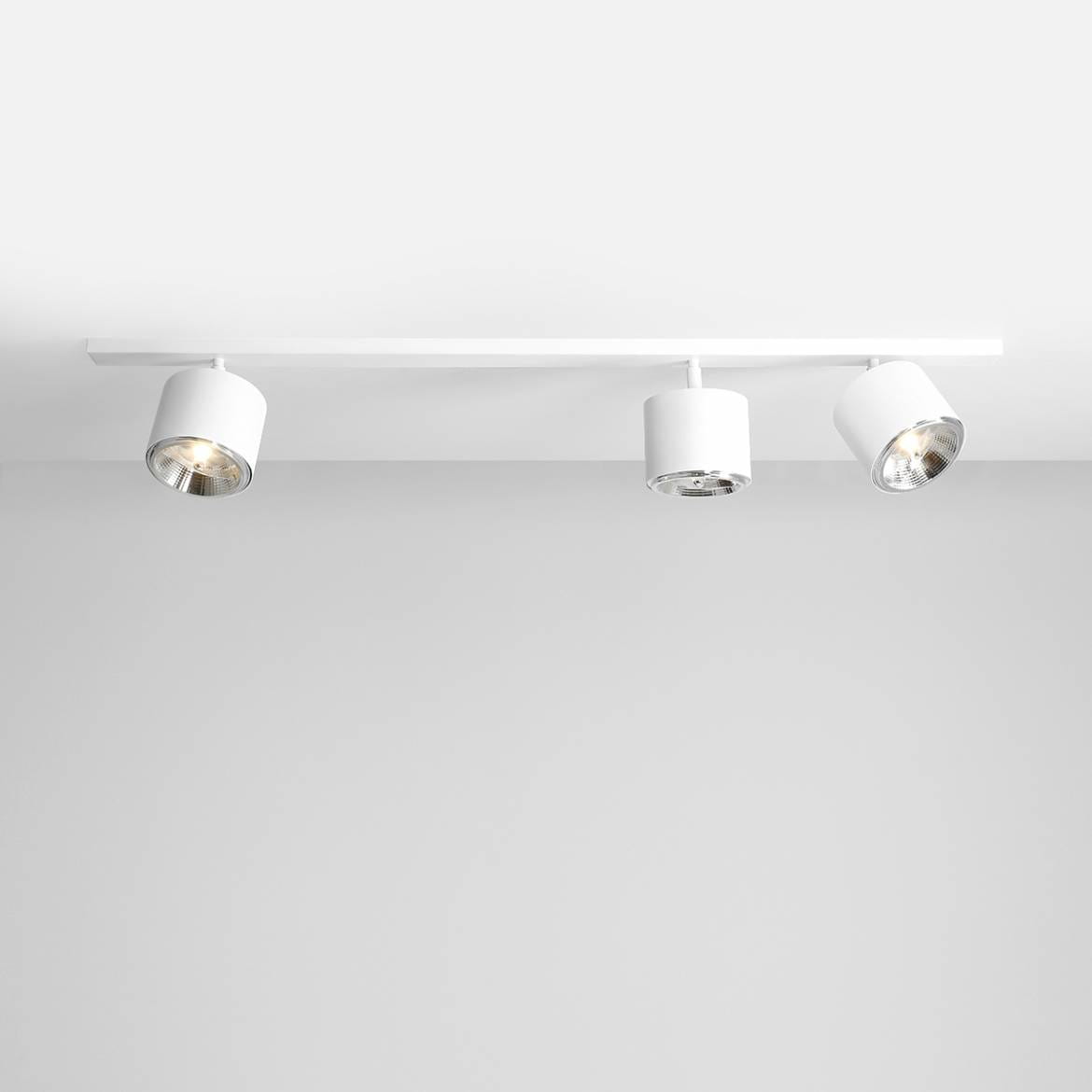Lampa sufitowa 3-punktowa w białym kolorze, z metalowymi kloszami, zamontowana na prostokątnej, białej listwie. Klosze mają cylindryczny kształt i emitują światło w różnych kierunkach. Tło jest jasne, co podkreśla design lampy.
