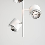 Lampa wisząca 3-punktowa w kolorze białym. Składa się z trzech reflektorów z metalicznymi kloszami, mocowanych na prostym, białym stelażu. Reflektory mają okrągły kształt i są skierowane w różne strony. Tło jasne, minimalistyczne.
