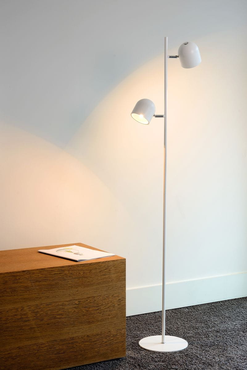 Biała lampa podłogowa Skanska z dwiema głowicami LED, umieszczona na okrągłej podstawie. Lampa stoi prosto obok drewnianego stolika, z widocznym białym tłem, emitując ciepłe światło. Kąt ujęcia frontalny, ukazujący detale konstrukcji.
