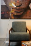 Lampa podłogowa Hester 1-punktowa w kolorze białym, z prostym, metalowym stelażem. Ujęcie z boku, lampka ustawiona obok zielonego fotela. Tło w stonowanych kolorach z abstrakcyjnym obrazem w odcieniach brązu i pomarańczu.