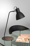 Czarna lampa biurkowa Shadi z jednopunktowym źródłem światła, z metalowym kloszem w kształcie stożka, umieszczona na cienkiej, giętej nodze i okrągłej podstawie. Lampa znajduje się na szklanym biurku obok dwóch otwartych broszur.