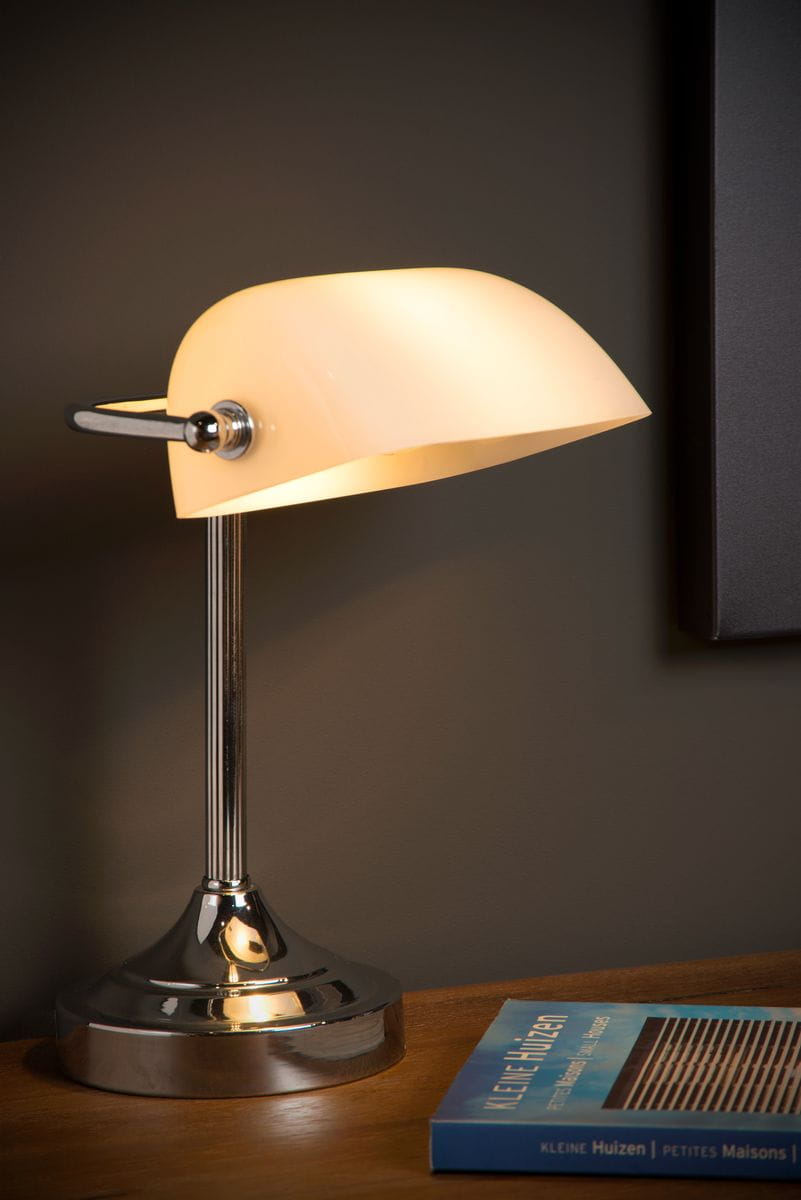Lampa biurkowa Banker 1-punktowa w kolorze białym z chromowanym podstawą. Posiada półokrągły klosz, lekko zaokrąglony, umieszczony na metalowej nóżce. Ujęcie z boku, na drewnianym biurku, obok książki. Tło w ciemnym kolorze.