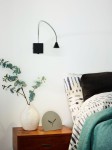 Kinkiet Buddy LED 1-punktowy w kolorze czarnym i chromowanym. Lampa ma elastyczny kabel, czarną czaszę oraz prostokątną bazę. Umieszczona na ścianie obok łóżka, w otoczeniu drewnianej szafki, wazonu z gałązkami oraz zegara.