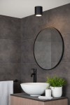 Czarna lampa sufitowa Aven IP65 1-punktowa, umieszczona w łazience, wisi na suficie. W tle widoczna okrągła lustro z czarną ramą, umywalka z białej ceramiki oraz drewniana szafka. Tło w odcieniach szarości, z teksturą kamienia.