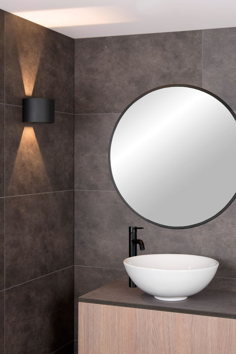 Czarny kinkiet Axi IP54 LED 1-punktowy, montowany na ścianie, emitujący światło w górę i w dół. Widoczny w nowoczesnej łazience, obok okrągłego lustra i umywalki na drewnianej szafce. Tło w neutralnych odcieniach szarości.