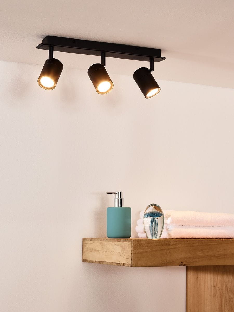 Czarna lampa sufitowa Lennert IP44 z trzema punktami świetlnymi, zamontowana na prostokątnej podstawie. Ujęcie z góry, w tle drewniana półka z niebieską butelką i ręcznikami. Jasne światło wydobywa się z lamp.