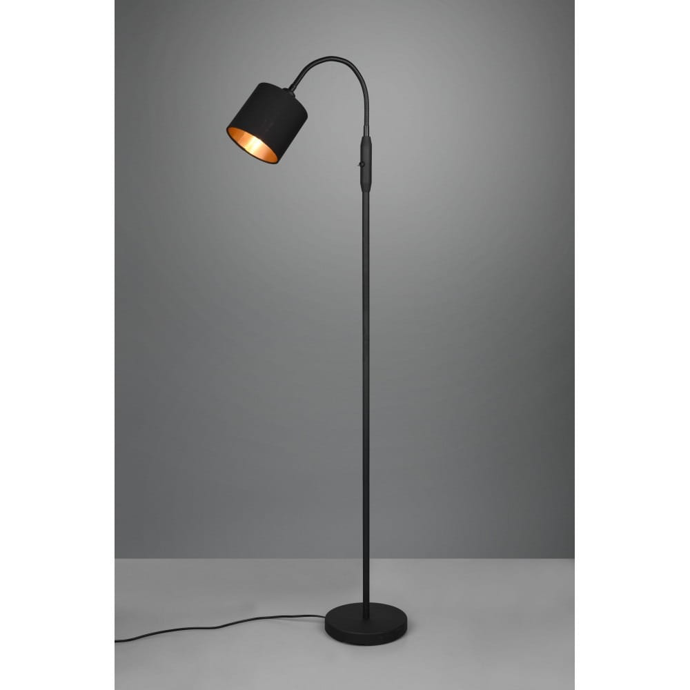 Lampa podłogowa Tommy w kolorze czarnym, z jednopunktowym źródłem światła. Posiada długą, prostą podstawę oraz zaokrągloną głowicę, która jest lekko nachylona. Kabel z włącznikiem widoczny obok podstawy. Tło w odcieniach szarości.