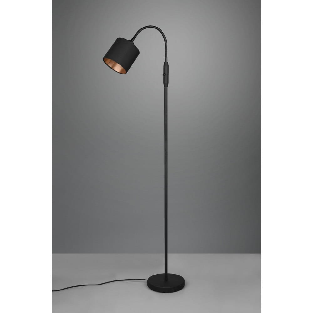 Czarna lampa podłogowa Tommy z jednopunktowym kloszem, o prostym, smukłym kształcie. Uchwyt łukowy i okrągła podstawa. Wnętrze klosza w kolorze miedzi. Lampa stoi na szarej powierzchni, w neutralnym tle.