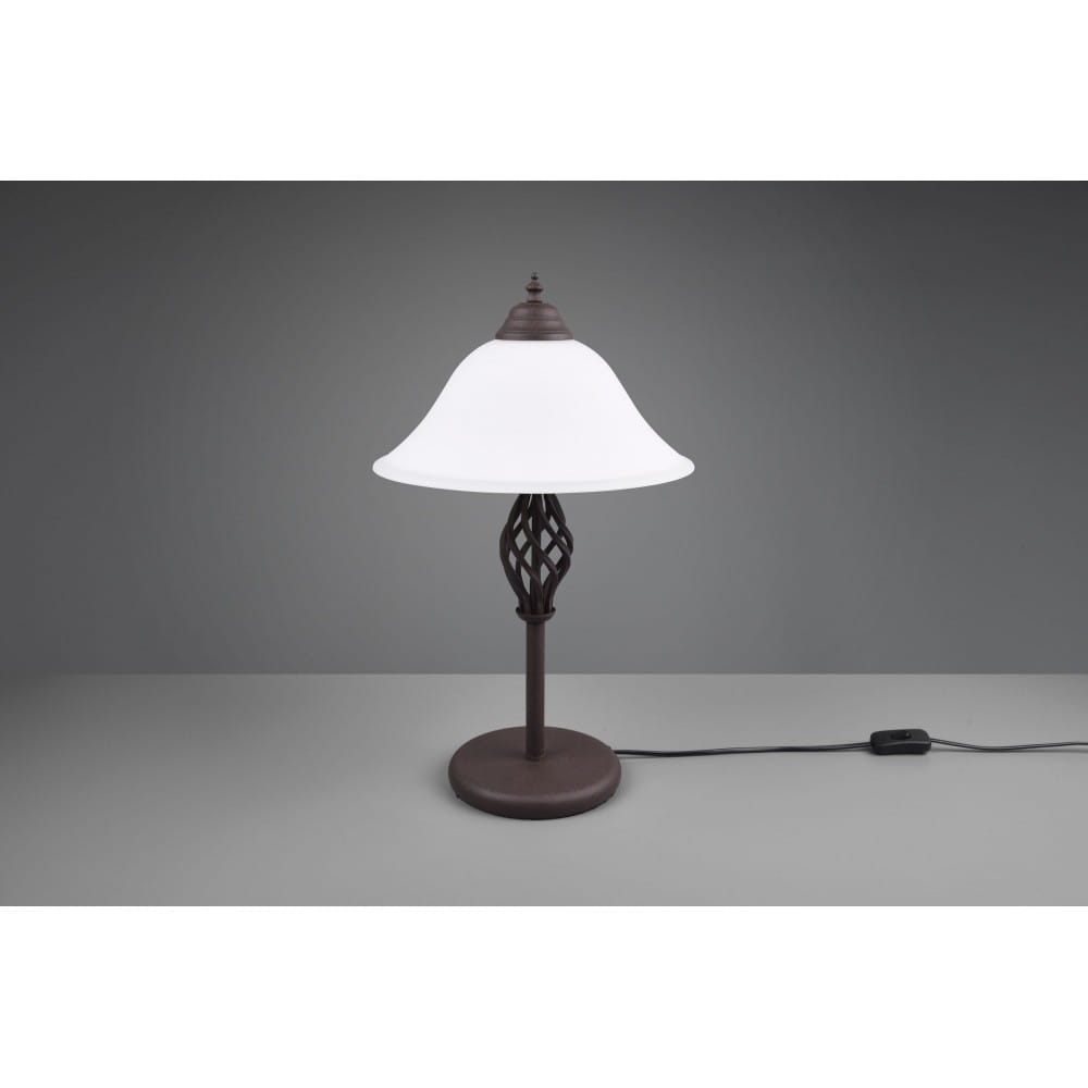 Lampa stołowa Rustica 2-punktowa w kolorze brązowym z białym kloszem. Kształt klosza przypominający dzwon oraz ozdobna, pleciona baza. Produkt umieszczony na szarej powierzchni, z widocznym przewodem z włącznikiem.