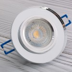 Okrągła oprawa sufitowa GU10 w transparentnym wykończeniu z metalowym brzegiem. Zbliżenie na wnętrze oprawy z soczewką, niebieskie mocowania do zabudowy. Produkt leży na jasnym tle z widocznym drewnianym wzorem.