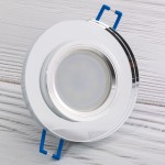 Okrągła oprawa do zabudowy GU10 w kolorze transparentnym, z metalicznym wykończeniem. Widoczny jest centralny otwór na żarówkę oraz niebieskie uchwyty montażowe. Produkt prezentowany z góry na jasnym tle.