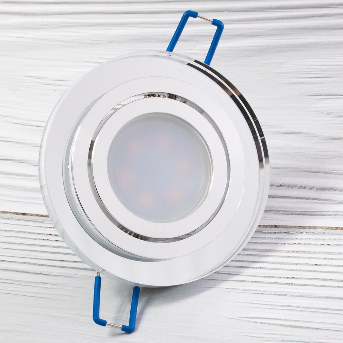 Okrągła oprawa oczko do zabudowy GU10, ruchoma, z transparentnym kloszem. Wykończenie w kolorze srebrnym, z widocznymi niebieskimi uchwytami mocującymi. Produkt leży na białym tle, ukazując szczegóły konstrukcji i designu.