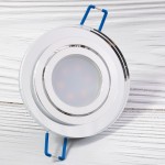 Okrągła oprawa oczko do zabudowy GU10, ruchoma, z transparentnym kloszem. Wykończenie w kolorze srebrnym, z widocznymi niebieskimi uchwytami mocującymi. Produkt leży na białym tle, ukazując szczegóły konstrukcji i designu.