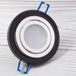 Okrągła, ruchoma oprawa oczko do zabudowy GU10 w kolorze czarnym z transparentnym kloszem. Widoczna srebrna ramka i niebieskie zaczepy montażowe. Produkt ułożony poziomo na jasnym tle.