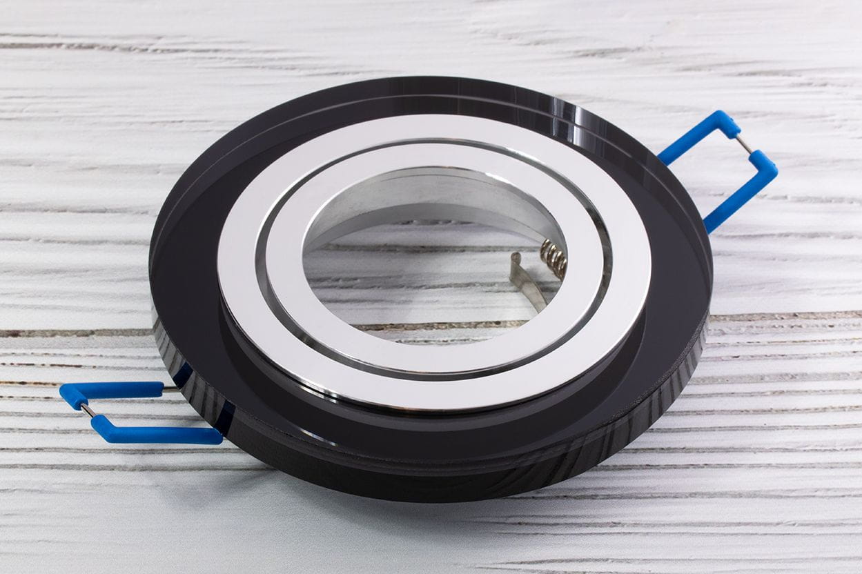 Oprawa oczko do zabudowy okrągła ruchoma GU10, w kolorze czarnym z transparentnym wykończeniem. Krawędź z polerowanego metalu, widoczne niebieskie uchwyty montażowe. Produkt leży na jasnym tle, ukazując detale konstrukcji.