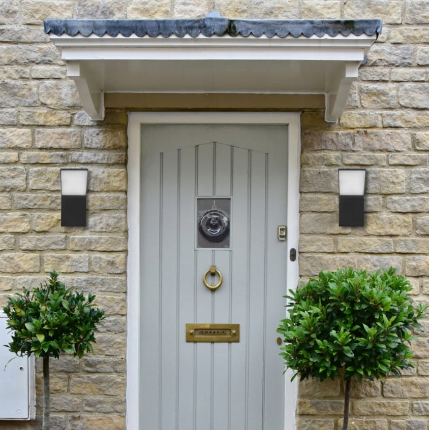 Kinkiet elewacyjny Clark LED w kolorze grafitowym, zamontowany obok drzwi. Ma prostokątny kształt, z białą, lekko przezroczystą osłoną u góry. Otoczenie to kamienna ściana oraz rośliny w donicach. Tło jasne, z wyraźnym kontrastem.