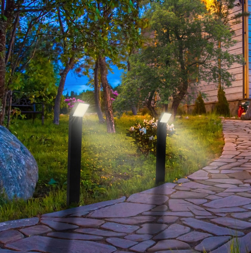 Grafitowa lampa stojąca Clark LED H80 w formie słupka, o prostokątnym kształcie, umieszczona na ścieżce w ogrodzie. Lampa emituje jasne światło, otoczona zielenią, kamieniami i kwiatami, w wieczornym świetle.