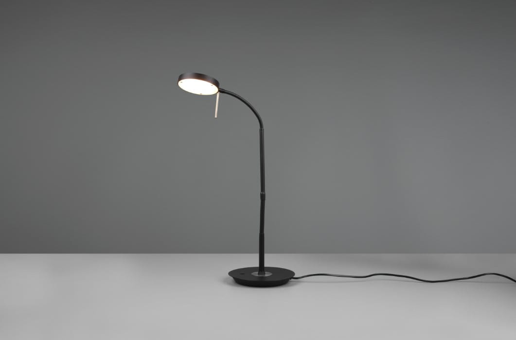 Czarna lampka biurkowa Monza LED z elastycznym ramieniem, podstawa okrągła. Ujęcie z boku, widoczna lampa z okrągłą głowicą emitującą światło. Kabel zasilający na stole, minimalistyczny design na szarym tle.