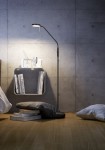 Czarna lampa podłogowa Monza LED z cienką, zakrzywioną nogą i okrągłą głowicą, ustawiona obok niskiego stolika z książkami. Tło stanowi surowa, betonowa ściana, a na podłodze leżą dwie poduszki.