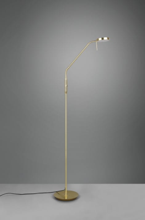 Monza LED lampa podłogowa w kolorze mosiądzu, o smukłej, wygiętej nodze i okrągłej podstawie. Ujęcie frontalne, lampa stoi na szarej powierzchni. Widoczny kabel zasilający.