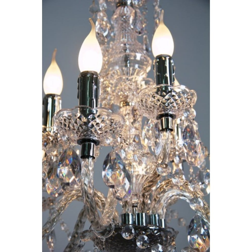 Luster żyrandol 9-punktowy, transparentny, z kryształowymi detalami i metalowymi elementami. Widoczne sześć świeczników z dekoracyjnymi kloszami, wiszące kryształy oraz elegancka podstawa. Ujęcie z bliska, ukazujące szczegóły konstrukcji.