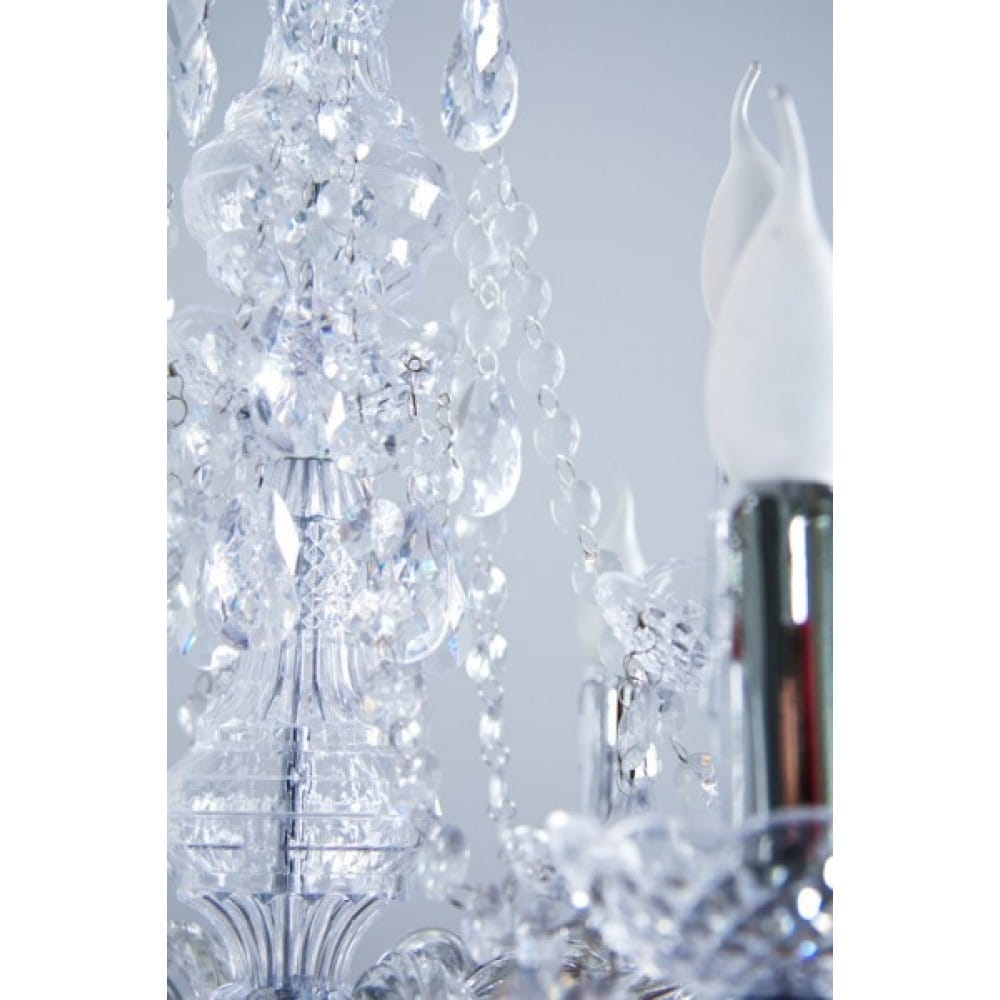 Luster żyrandol 9-punktowy z przeźroczystymi kryształami, dekorowany wiszącymi łańcuchami i efektownymi detalami. Widoczne białe klosze na świeczki oraz chromowane mocowanie. Ujęcie zbliżeniowe na górną część lampy.