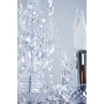 Luster żyrandol 9-punktowy z przeźroczystymi kryształami, dekorowany wiszącymi łańcuchami i efektownymi detalami. Widoczne białe klosze na świeczki oraz chromowane mocowanie. Ujęcie zbliżeniowe na górną część lampy.