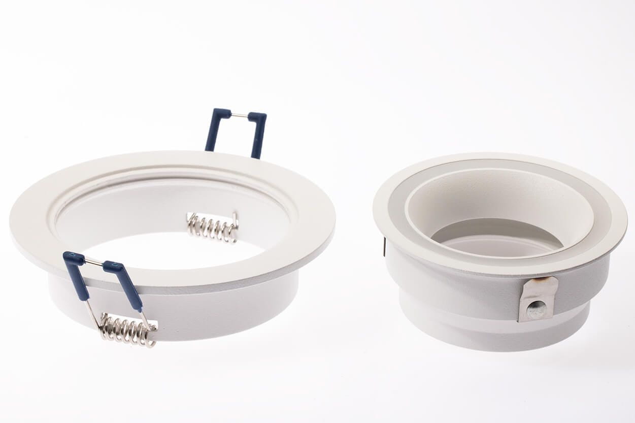 Biała oprawa oczko do zabudowy okrągła GU10, z widocznymi sprężynami mocującymi, w układzie stojącym. Produkt składa się z dwóch części; jedna z nich ma metalowe mocowanie. Gładka powierzchnia, prosty design, na jednolitym tle.