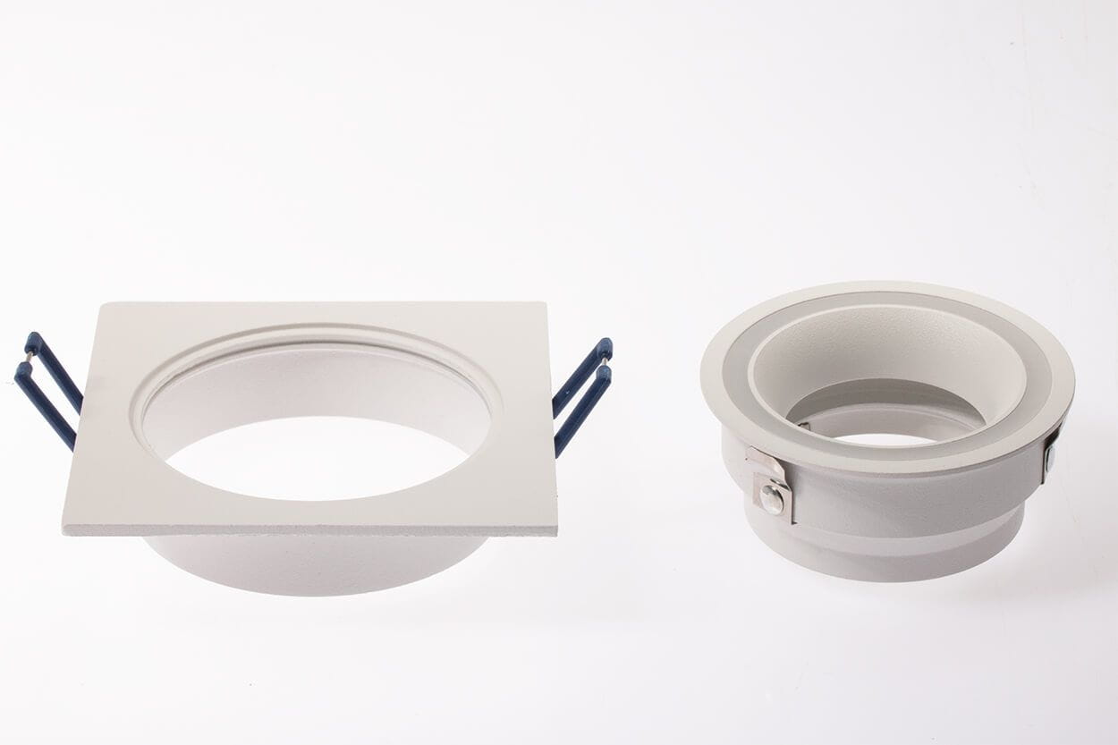 Kwadratowa oprawa oczko do zabudowy GU10 w kolorze białym, z widocznymi metalowymi uchwytami. Produkt leży na białym tle, zbliżenie na detal. W zestawie znajduje się również okrągła oprawa o gładkiej fakturze.