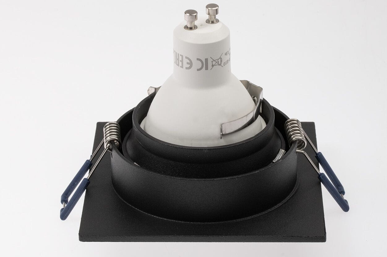 Kwadratowa oprawa oczko do zabudowy w kolorze czarnym, przeznaczona do żarówek GU10. Widok z góry, pokazujący metalowe sprężyny mocujące oraz białą część z gwintem. Prosta, nowoczesna konstrukcja. Tło białe.