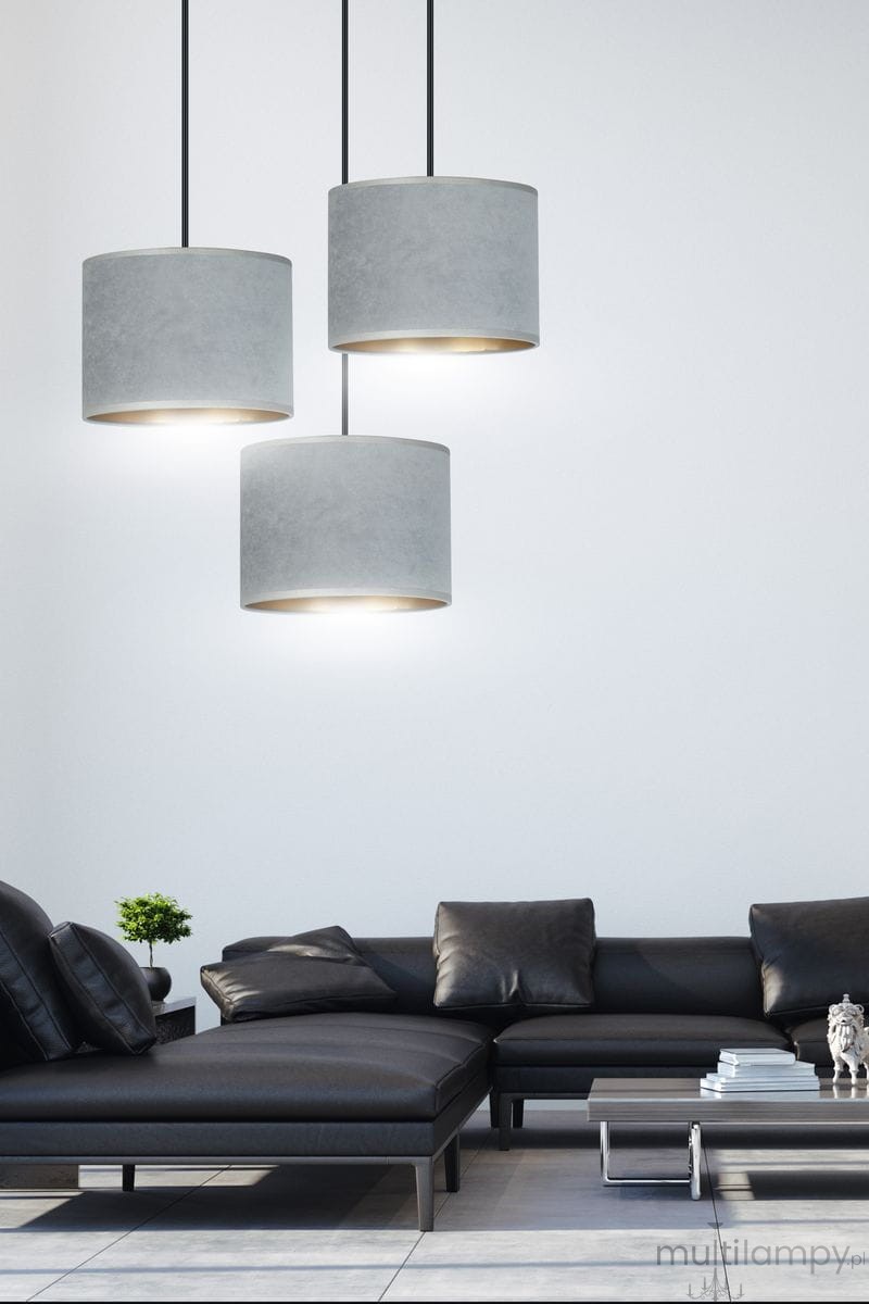 Lampa wisząca Hilde 3-punktowa w odcieniu szarym, składająca się z trzech cylindrycznych kloszy z materiału, zawieszona na czarnych linkach. Widoczna w nowoczesnym wnętrzu z czarną sofą i niskim stolikiem.