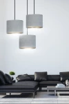 Lampa wisząca Hilde 3-punktowa w odcieniu szarym, składająca się z trzech cylindrycznych kloszy z materiału, zawieszona na czarnych linkach. Widoczna w nowoczesnym wnętrzu z czarną sofą i niskim stolikiem.