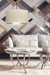 Lampa wisząca Hilde 1-punktowa w kolorze białym, z okrągłym kloszem wykonanym z materiału. Zawieszona nad sofą w jasnym wnętrzu, na tle wielokolorowej ściany z desek. Widoczne stoliki kawowe z metalowymi nóżkami.