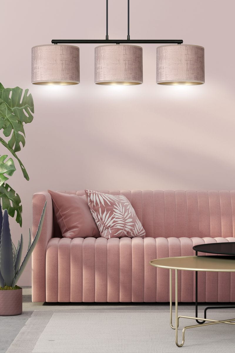 Lampa wisząca Hilde 3-punktowa w odcieniu różowym, z okrągłymi kloszami z materiału, zawieszona na czarnej konstrukcji. W tle różowa ściana, sofka z teksturowanym obiciem oraz dekoracyjne poduszki. W otoczeniu roślinności.