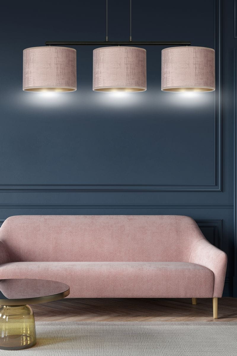 Lampa wisząca Hilde 3-punktowa w kolorze różowym z trzema cylindrycznymi kloszami na czarnym stelażu. Klosze mają delikatną fakturę, a lampa zawieszona jest nad różową sofą w nowoczesnym wnętrzu z ciemnoniebieską ścianą.