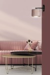 Kinkiet Hilde w kolorze różowym, z metalowym czarnym mocowaniem, umieszczony na ścianie. Lampa ma cylindryczny klosz o teksturowanej powierzchni. W tle widoczna różowa sofa oraz czarne i złote stoliki kawowe.