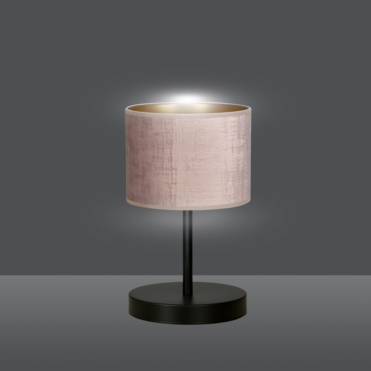 Lampa stołowa Hilde w kolorze różowym, z cylindrycznym abażurem o matowej fakturze, na czarnej podstawie. Ujęcie frontalne, lampa emituje delikatne światło, tworząc przytulną atmosferę. Idealna do wnętrz.