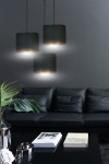 Czarna lampa wisząca Hilde z trzema cylindrycznymi kloszami, zawieszona na cienkich przewodach. Klosze mają jasne wnętrze i emitują delikatne światło, umieszczone nad czarną sofą w nowoczesnym wnętrzu. W tle widać roślinę oraz książki na stole.