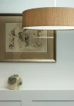 Lampa wisząca Turda D65 w kolorze białym i złotym, z okrągłym abażurem o fakturze tkaniny, zawieszona na cienkim kablu. W tle widoczna ramka z grafiką oraz ceramiczna dekoracja w kształcie głowy.
