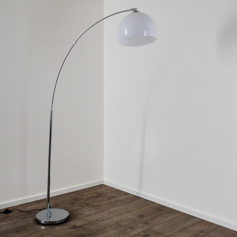 Lampa podłogowa Brasilia 1-punktowa z chromowanym stelażem i białą, półkulistą kloszem. Ujęcie z boku, lampa ma smukłą, zakrzywioną nogę i okrągłą podstawę. Tło jasne, jednolite, podłoga drewniana.