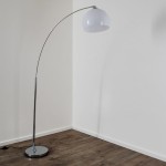 Lampa podłogowa Brasilia 1-punktowa z chromowanym stelażem i białą, półkulistą kloszem. Ujęcie z boku, lampa ma smukłą, zakrzywioną nogę i okrągłą podstawę. Tło jasne, jednolite, podłoga drewniana.