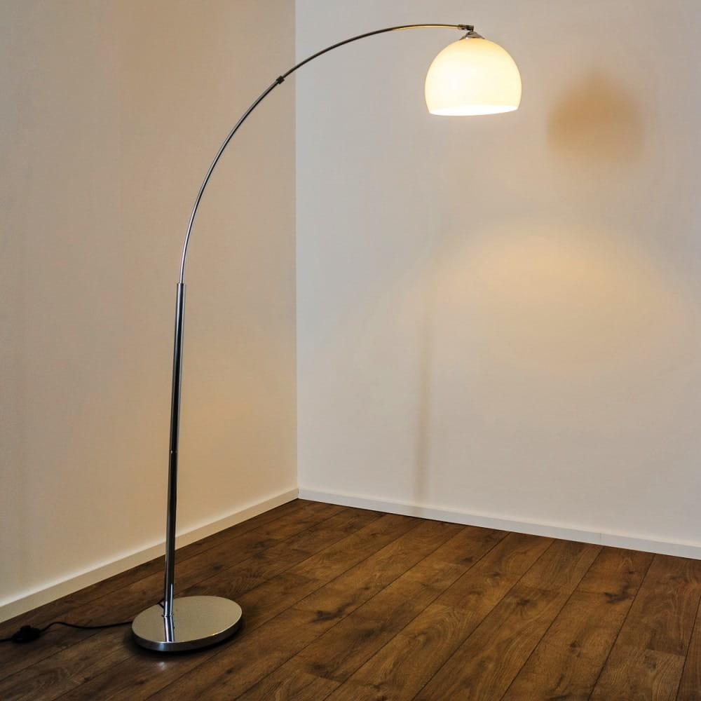 Lampa podłogowa Brasilia 1-punktowa w chromowanej obudowie, z okrągłym, białym kloszem. Ujęcie z boku pokazuje elegancki, zakrzywiony kształt i stabilną podstawę. Produkt umieszczony na drewnianej podłodze, w tle jasna ściana.