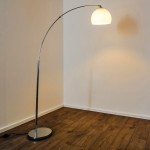 Lampa podłogowa Brasilia 1-punktowa w chromowanej obudowie, z okrągłym, białym kloszem. Ujęcie z boku pokazuje elegancki, zakrzywiony kształt i stabilną podstawę. Produkt umieszczony na drewnianej podłodze, w tle jasna ściana.