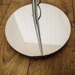 Lampa podłogowa Brasilia z chromowanym stelażem, widok z dołu. Okrągła, lustrzana podstawa odbijająca światło, z cienką, prostą nogą. Tło to drewniana podłoga w ciepłym odcieniu.